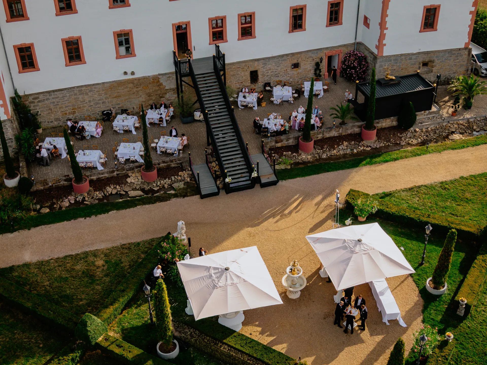 Luftaufnahme der Terrasse mit weiß gedeckten Tischen, Freitreppe und Sonnenschirmen am Brunnen