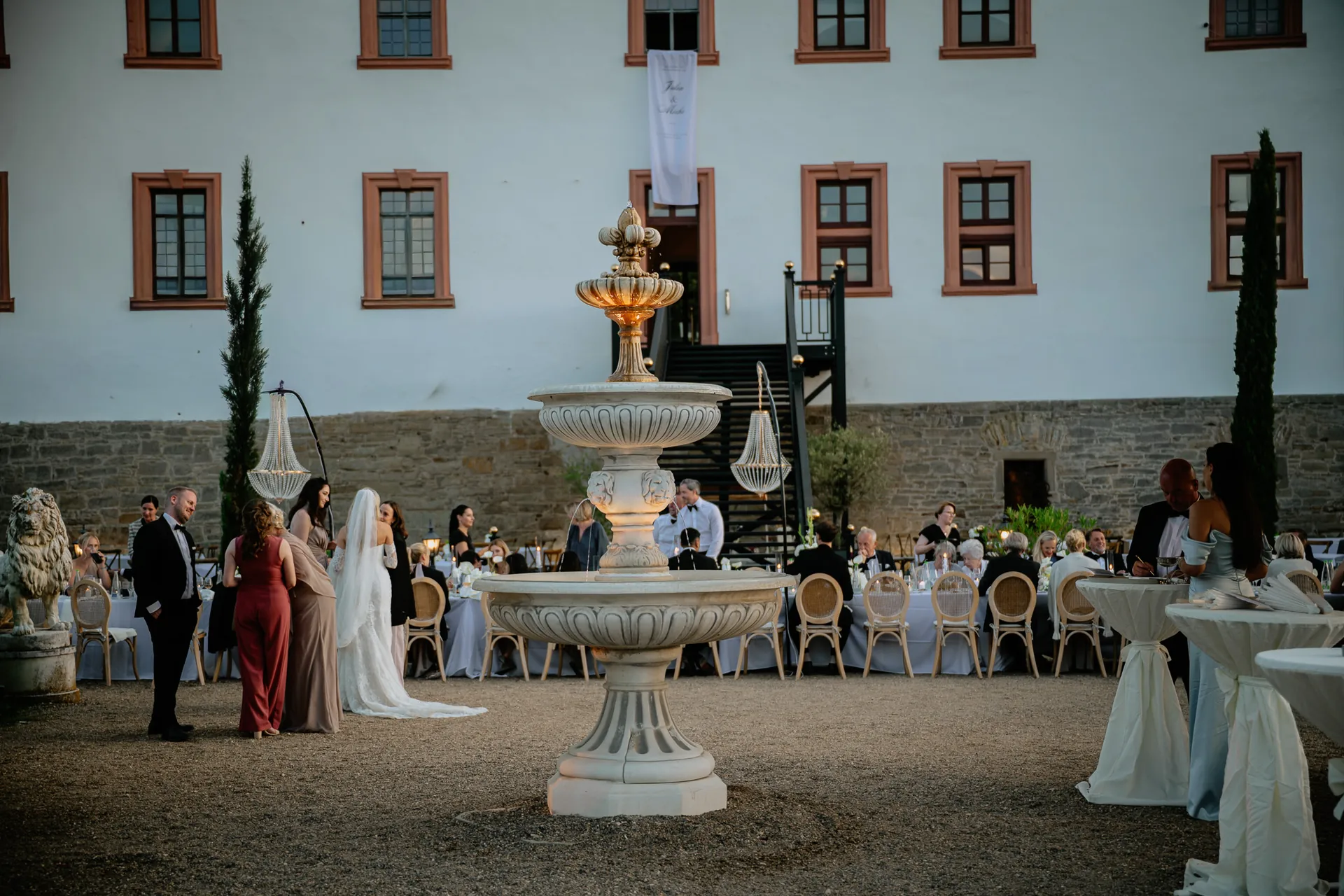 Gäste an langen Tafeln rund um den beleuchteten Brunnen vor der Schlossfassade in der Abenddämmerung