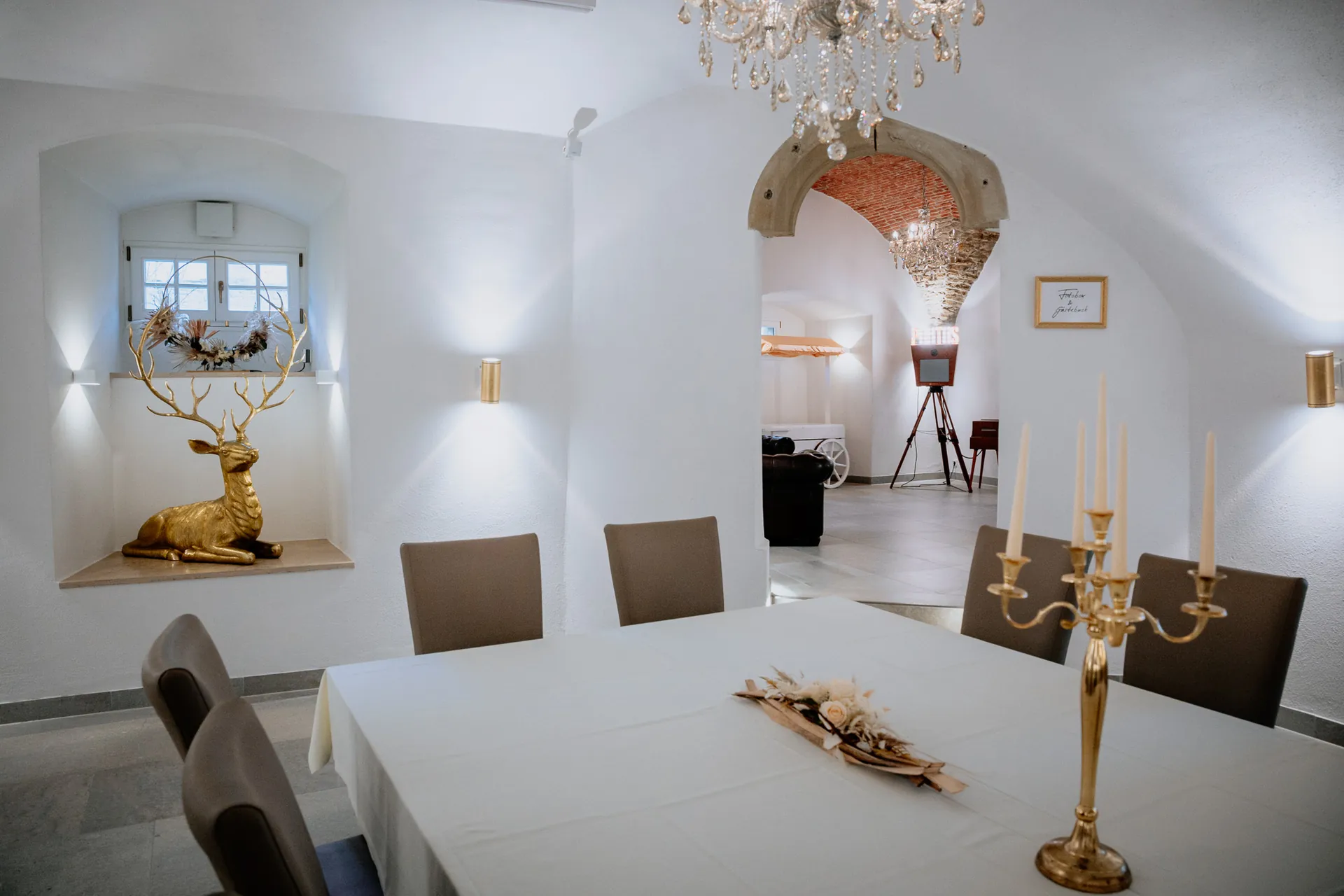 Gewölbesaal mit weiß gedeckter Tafel, Kristallleuchter, goldenem Hirsch in der Fensternische und Blick ins Foyer